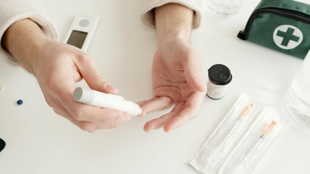 IMAGEM DE UM HOMEM, FAZENDO USO DO APARELHO DE GLICOSIMETROU OU MEDIDOR DOS NIVEIS DE GLICEMIA CAPILAR, MOSTRANDO APENAS AS MÃO COM UM EXEMPLO DE ESTA FURANDO COM A CANETA DO APARELHO, NA MESA TEMOS 2 SERINGA DE INSULINA, E OS ACESSORIOS DO APARELHO DE GLICOSE